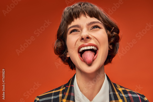 Playful female student in college uniform sticking out tongue, lively on orange background
