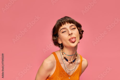 cheeky carefree girl in 20s with short brunette hair sticking tongue out on pink background