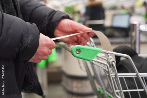 An unrecognizable man's hands hold a sales receipt, next to a grocery cart in a supermarket.