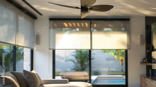 a living room.the rolling shade in the back of it in a insteresting way. the ceiling fan in the foreground, and the window with a smar roller shade in the background.