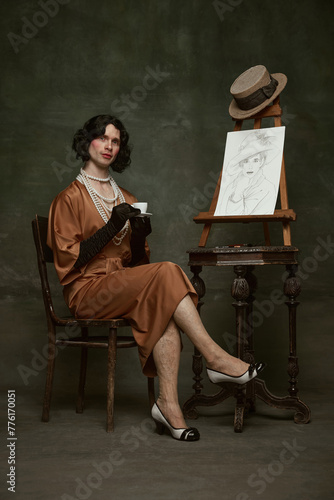 Fototapeta Elegant man in silky, old-fashion dress sitting by easel with self-portrait and drinking coffee against vintage studio background