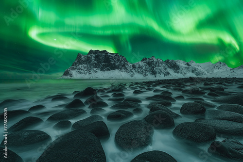 Low angle view of waves flowing over the rocks in the foreground with snow-capped mountains in the background at night. Aurora Borealis dancing over the beautiful rocky coastline of Uttakleiv beach