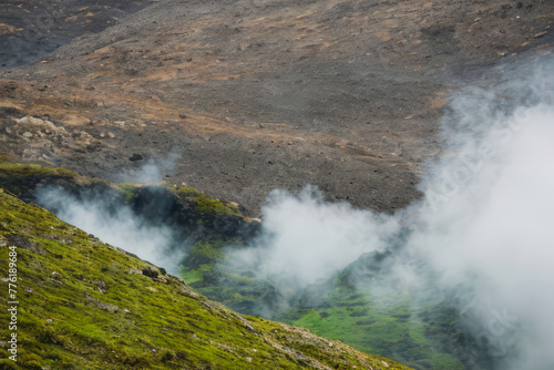 A cascading waterfall plunges down moss-covered mountains in a geothermal landscape