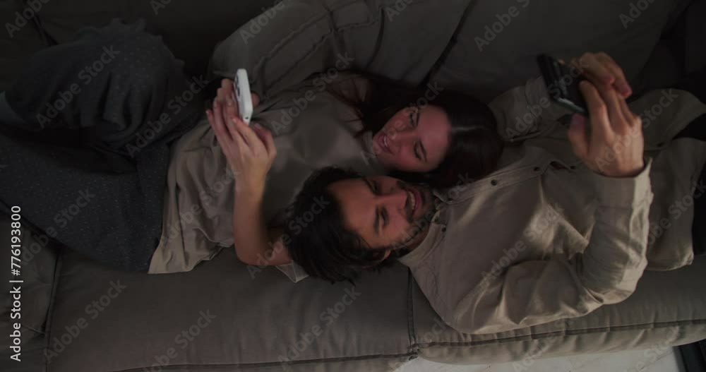 A brunette guy with stubble and a girl in a beige T-shirt lie head to head on the sofa and watch something on their smartphones during their evening relaxation on the sofa A brunette guy with stubble and a girl in a beige T-shirt lie head to head on the sofa and watch something on their smartphones during their evening relaxation on the sofa