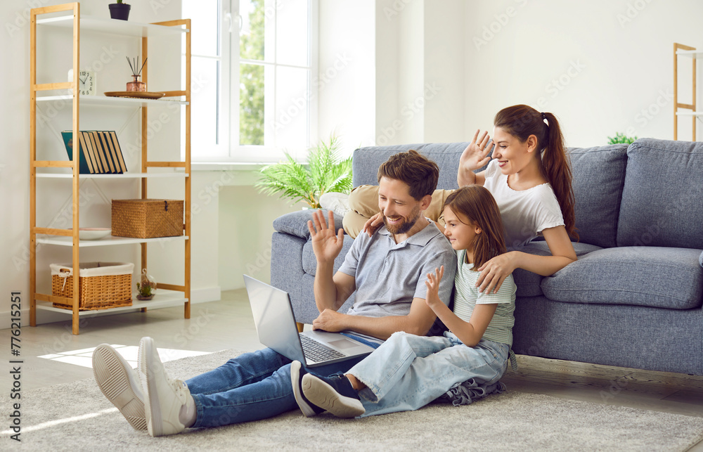Happy family using laptop and video calling relatives. Young mother, father and child daughter sitting in living room at home, looking at screen of modern notebook computer, waving hello and smiling Happy family using laptop and video calling relatives. Young mother, father and child daughter sitting in living room at home, looking at screen of modern notebook computer, waving hello and smiling