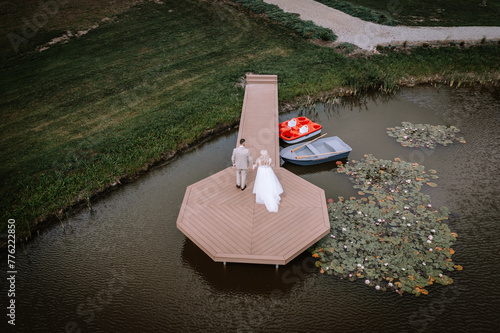 Valmiera, Latvia - Augist 13, 2023 - A bride and groom walk on a narrow pier leading to an octagonal deck on a pond, with a red paddle boat and a gray rowboat nearby. photo