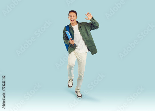 Happy school, college or university student hurries for class, sees friend and says hello. Full body front view portrait smiling young man teen boy with book walks on light blue background waving hand