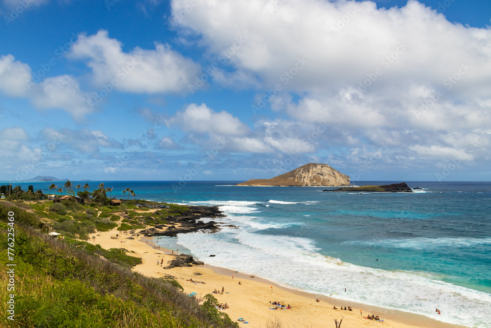 a beautiful spring landscape along the coast of Oahu with a sandy beach, blue ocean water, people relaxing, lush green trees and plants, rocks, blue sky and clouds in Honolulu Hawaii USA