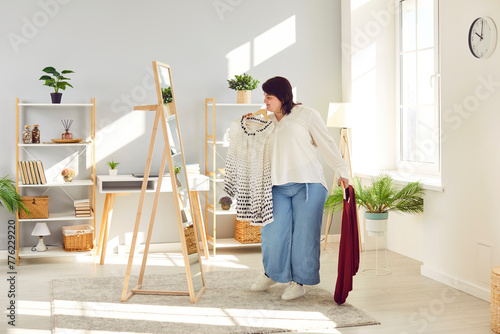 Overweight woman looking at mirror trying to choose clothes at home wardrobe. Plus size young woman holding hangers with blouses, thinking what to wear. Body positive concept photo
