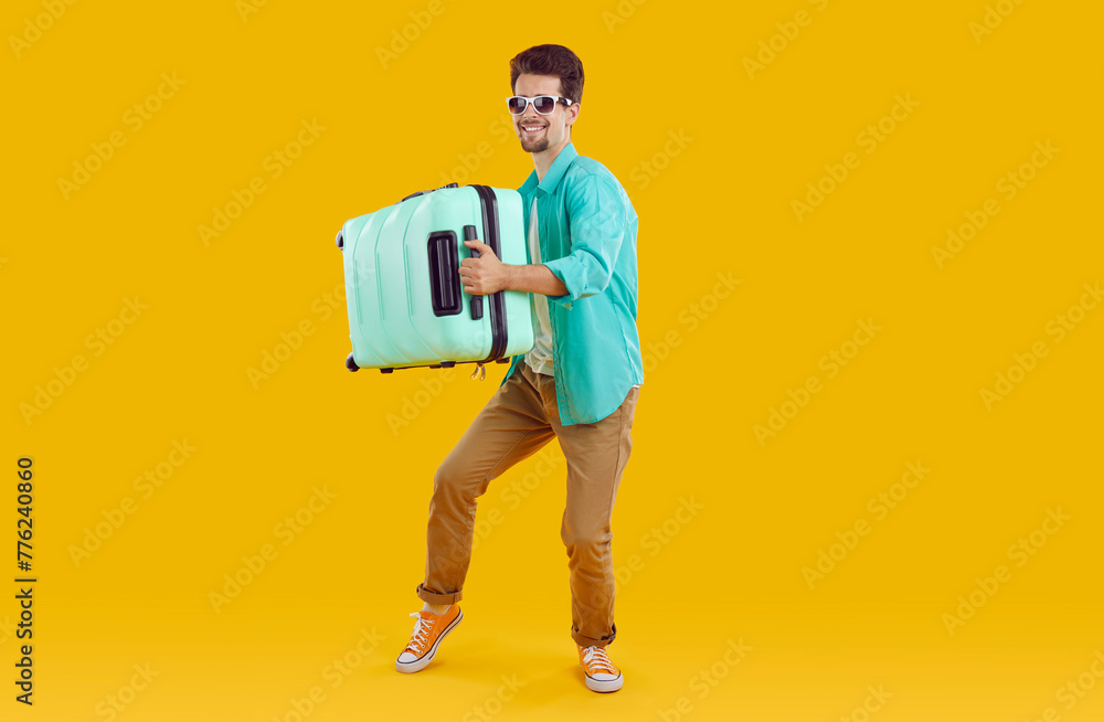 Happy young man traveler in sunglasses carrying suitcase. Cheerful handsome man wearing casual clothes standing with luggage at copy space on yellow background ready for vacation