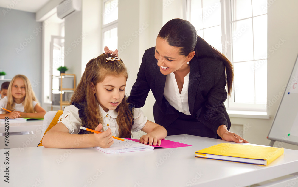 Education, teaching and knowledge. Caring teacher in classroom during lesson helps little schoolgirl to complete school assignment. Little girl is sitting at desk with notebook in which she writes. Education, teaching and knowledge. Caring teacher in classroom during lesson helps little schoolgirl to complete school assignment. Little girl is sitting at desk with notebook in which she writes.