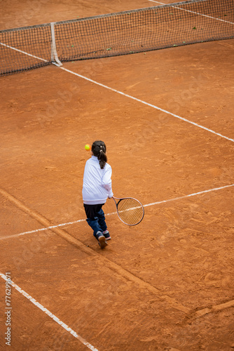 A little girl plays tennis on a red clay court. She runs and trains to learn the beautiful sport of tennis. Competitiveness, athlete, sporty child.