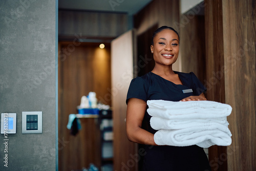 Happy black chambermaid with stack of clean towel in a hotel room looking at camera.