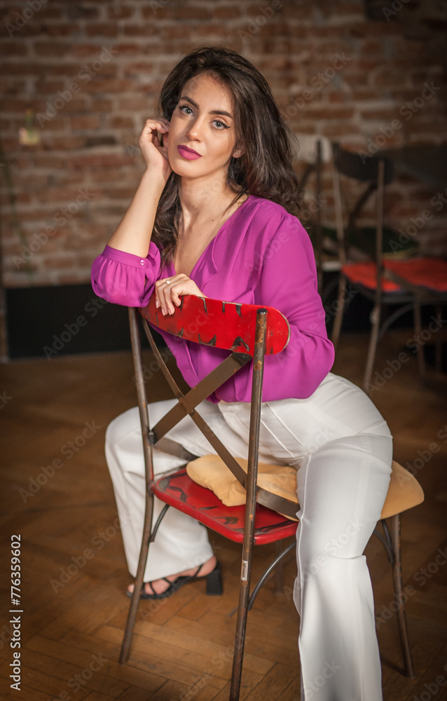 Beautiful young brunette woman in a purple blouse and white pants posing indoor against a brick wall. Portrait of a beautiful young brunette woman
