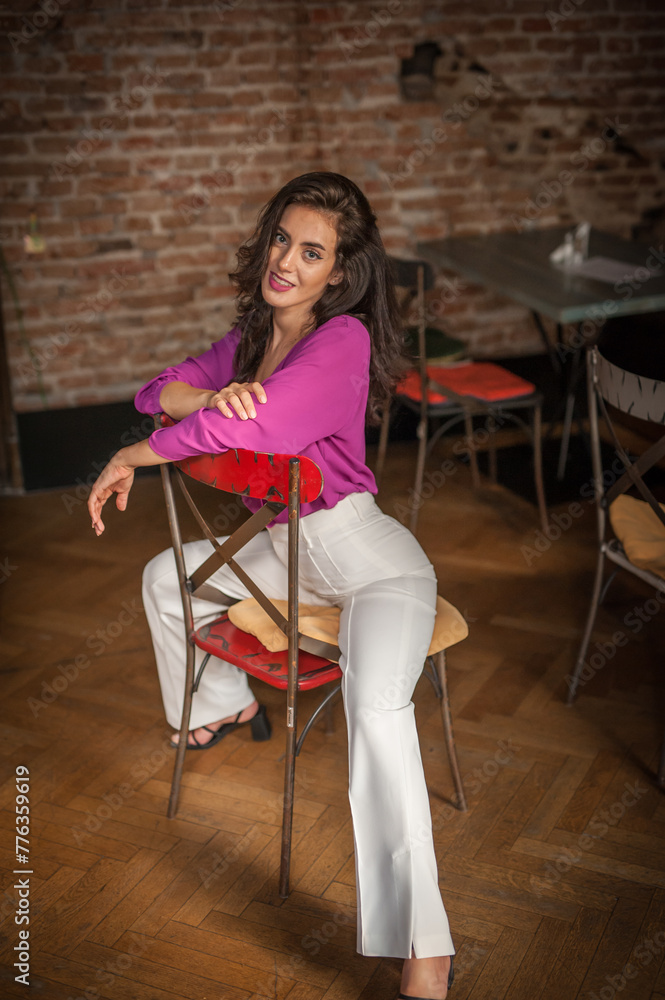 Beautiful young brunette woman in a purple blouse and white pants posing indoor against a brick wall. Portrait of a beautiful young brunette woman