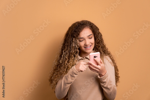 happy female student smiling and holding mobile phone in beige studio background. communication, app, connection concept.