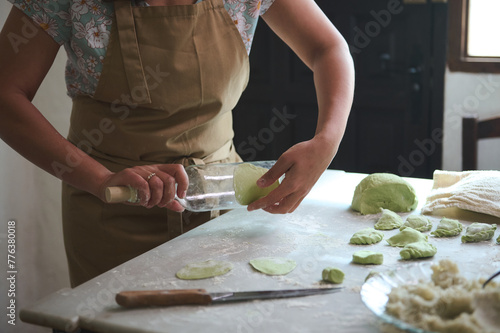 Close-up housewife using glass wine bottle as rolling pin, rolling out green spinach dough for making dumplings, standing at kitchen table sprinkled with flour in the rural house kitchen