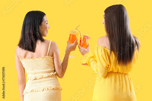 Beautiful young women with glasses of tasty aperol spritz cocktail on yellow background, back view