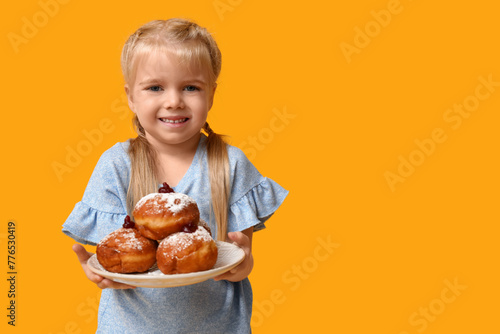 Cute happy little girl holding plate with tasty donuts on yellow background. Hanukkah celebration