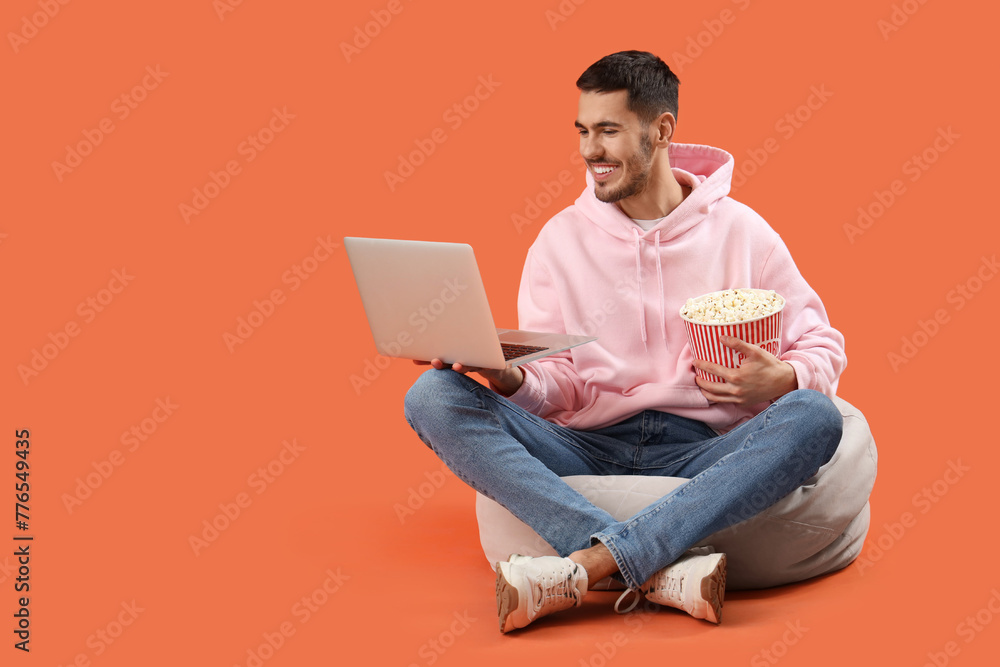 Handsome young happy man with laptop and popcorn resting on beanbag chair against orange background Handsome young happy man with laptop and popcorn resting on beanbag chair against orange background