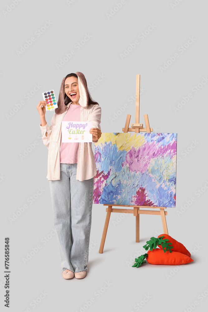 Εκτύπωση καμβά Happy female artist in bunny ears with Easter greeting card, toy carrots and eas