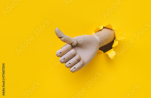 A wooden hand protrudes from a torn hole in yellow paper and shows a big thumbs up like gesture. The concept of approval and that everything will be fine. Artificial intelligence, robot.
