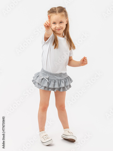 The girl happily and expressively points her finger at something in a T-shirt and skirt, standing on a white background. Photo.