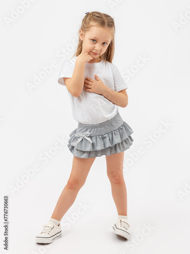 Timidity, indecision. A 5-year-old girl poses in a white T-shirt and a gray skirt on a white background. Hands at the face. Photo. Copy space.