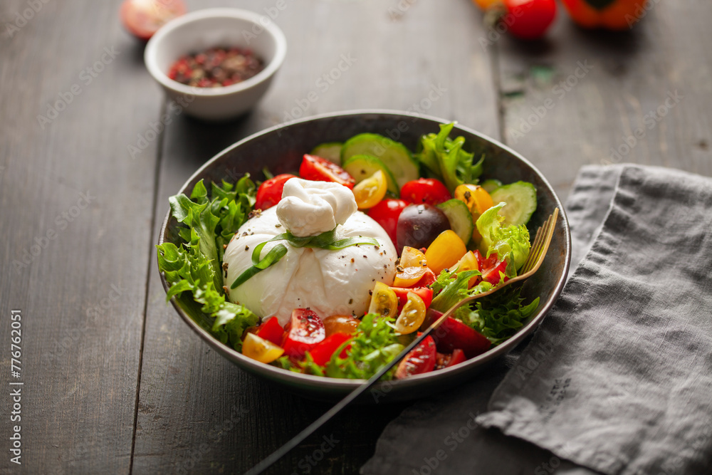 Salad with traditional italian burrata cheese. Burrata ball with cherry tomatoes, cucumber slices, lettuce  and peppercorn in bowl on the table
