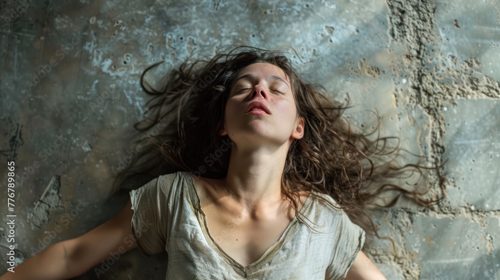 Naklejka premium A young woman lies back with eyes closed, displaying a look of tranquility against a grunge, textured concrete backdrop, her hair spread out.
