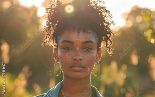 A woman with curly hair and hoop earrings is standing in a field. The sun is shining on her face, creating a warm and inviting atmosphere © imagineRbc