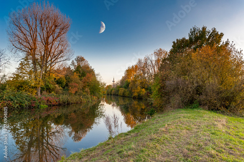 Herbststimmung am Fluß Nidda in Frankfurt am Main mit bunt verfärbten Laubbäumen und Sträuchern am Ufer und dem aufgehenden  Halbmond am Horizont