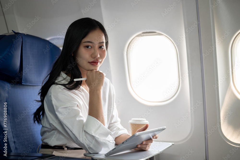 Fototapeta premium A confident Asian businesswoman is sitting at a window seat on a plane with her digital tablet.