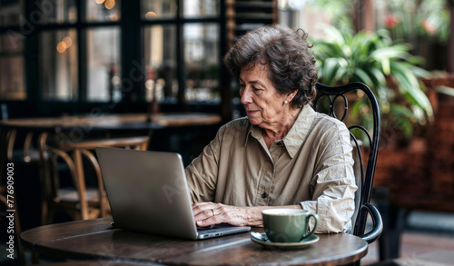 An elderly woman of 60  70 years old is sitting in a modern cafe with a laptop. Senior happy business woman using pc technologies, working, chatting, spending time in social media internet, ecommerce. photo