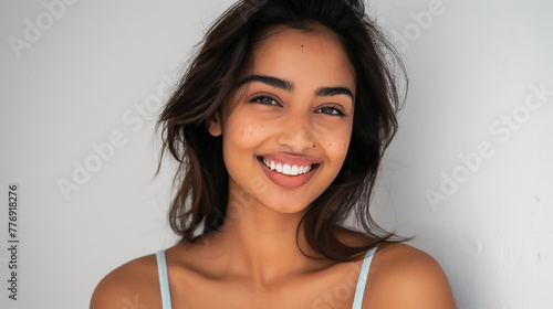 A beautiful Indian young woman with dark hair wearing a light blue tank top smiling and posing for the camera on a white background