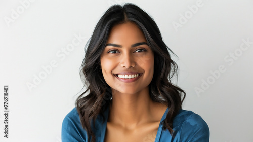 A beautiful Indian woman with dark hair wearing a blue top smiling against a white background in a studio shot photo