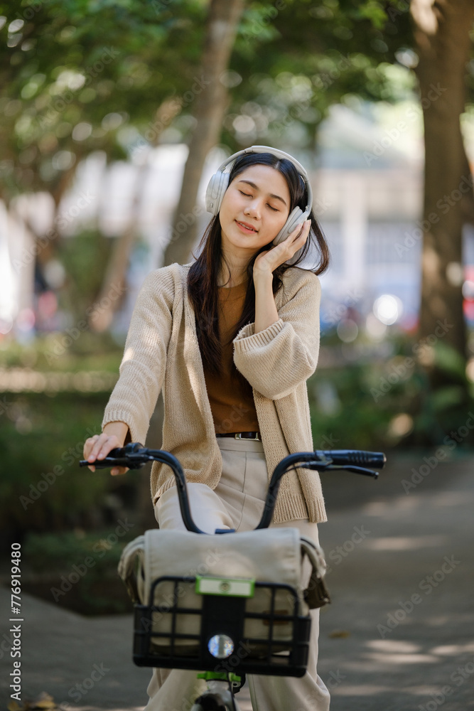 Fototapeta premium Young asian woman using headphones while riding bicycle in city park