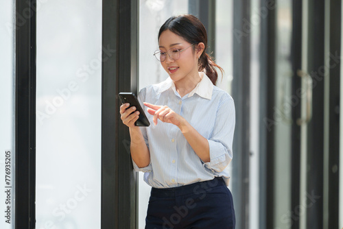 Happy Asian businesswoman in glasses standing smiling looking at data Talk on the phone using your smartphone for communication. Check through the application Go online while in the modern office.