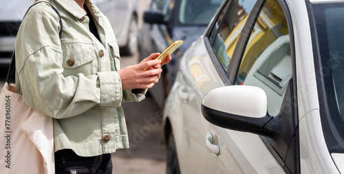 The woman unlocks the car rental vehicle using the mobile app. Woman with mobile phone connecting parked car on street.