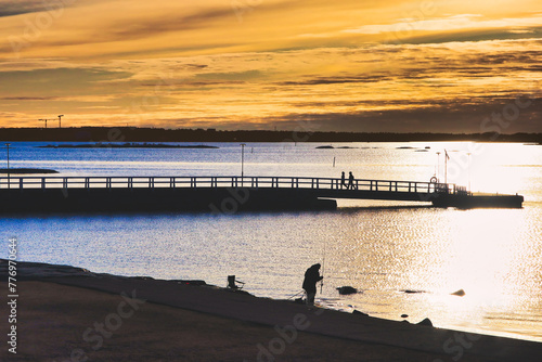 Landscape with a sea fishing fisherman on the waterfront and two walkers on the pier in the light of the golden hour. People seen in distance, in silhouette, not recognizable.