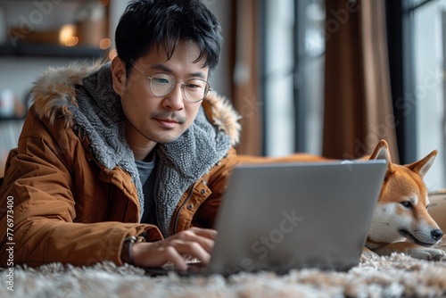 A man is sitting on the floor with his companion dog, a carnivore breed, sharing a happy moment while using a laptop. The scene is set against a backdrop of a curtain and wood