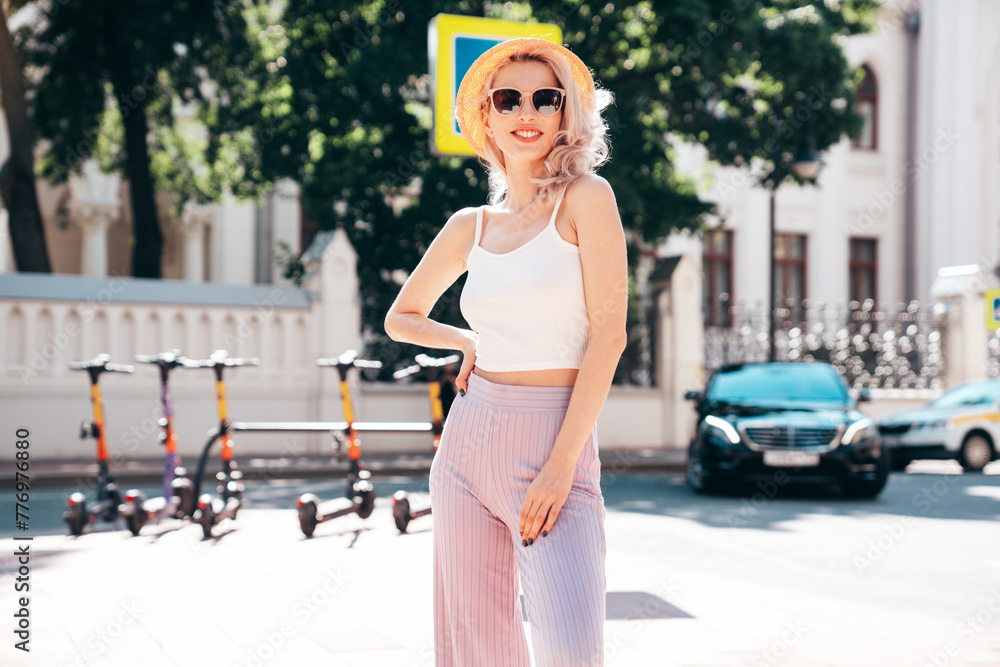 Young beautiful smiling blond hipster woman in trendy summer clothes. Carefree female posing in the street at sunny day. Positive model outdoors at sunset. Cheerful and happy in hat, sunglasses