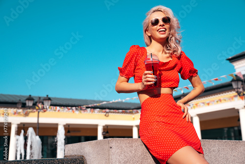 Young beautiful smiling hipster woman in trendy summer clothes. Carefree model posing in the street. Holding and drinking fresh vegetable cocktail smoothie drink in plastic cup with straw. Sunglasses