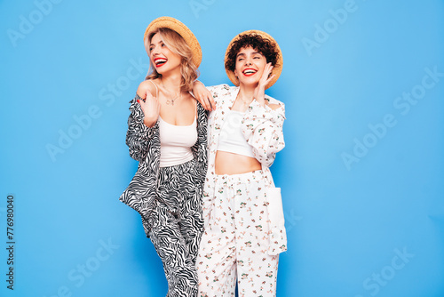 Two young beautiful smiling brunette hipster female in trendy summer costume clothes. Sexy women posing near blue wall in studio. Positive models having fun. Cheerful and happy. In hat