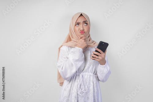 Thoughtful young Asian Muslim woman wearing white dress and hijab, using smartphone while holding her chin and thinking with serious expression over isolated white background