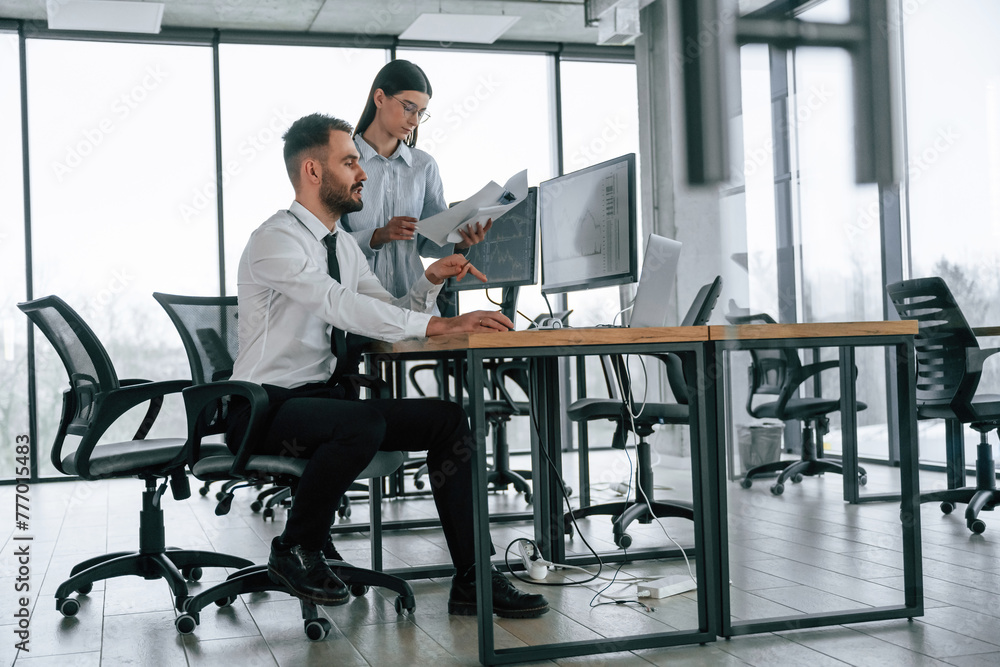 Woman is standing and holding documents. Two employees are working in the office together Woman is standing and holding documents. Two employees are working in the office together
