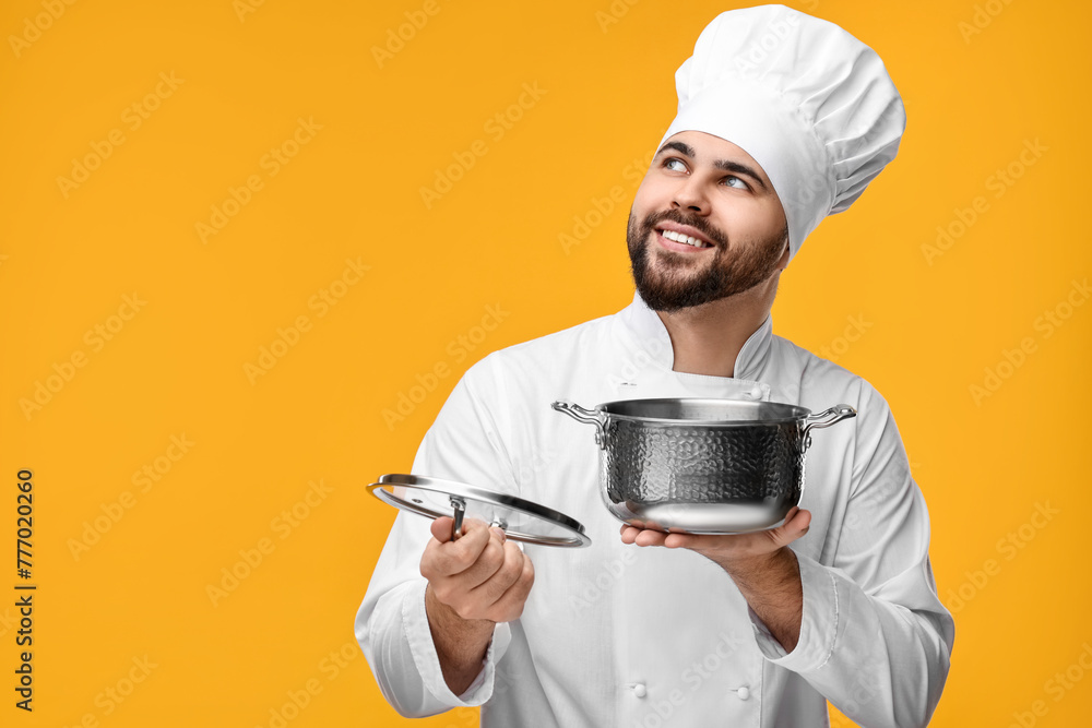 Happy young chef in uniform holding cooking pot on orange background. Space for text Happy young chef in uniform holding cooking pot on orange background. Space for text