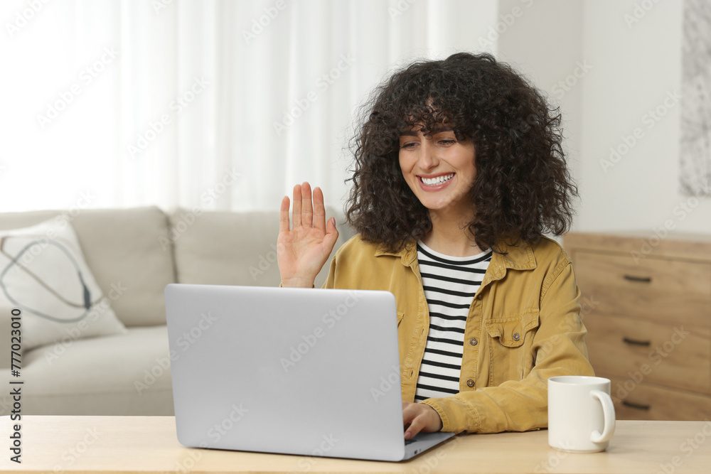 Happy woman waving hello during video call at table in room Happy woman waving hello during video call at table in room