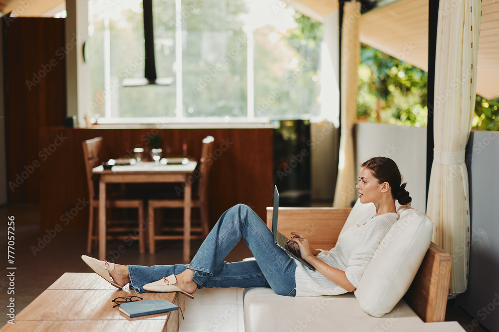 Woman enjoying a relaxing weekend at home, sitting on a modern sofa in her cozy living room With a smile on her face, she chats online, does some online shopping, and works on her laptop Surrounded by