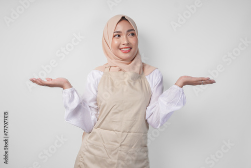Excited young Asian Muslim woman in hijab and cream apron pointing to the empty copy space on the left and right side. Ramadan advertisement concept